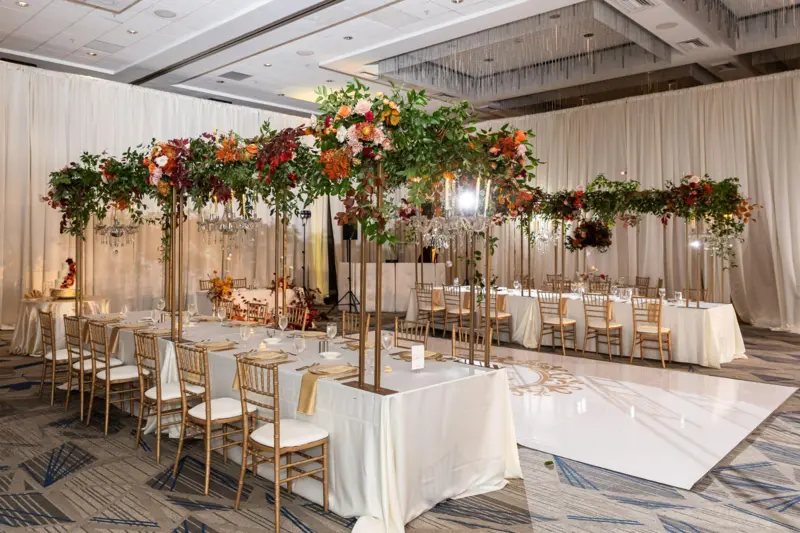 Ballroom reception with long banquet tables under a custom metal canopy frame covered in greenery and flowers, surrounded by gold chairs and a white dance floor.