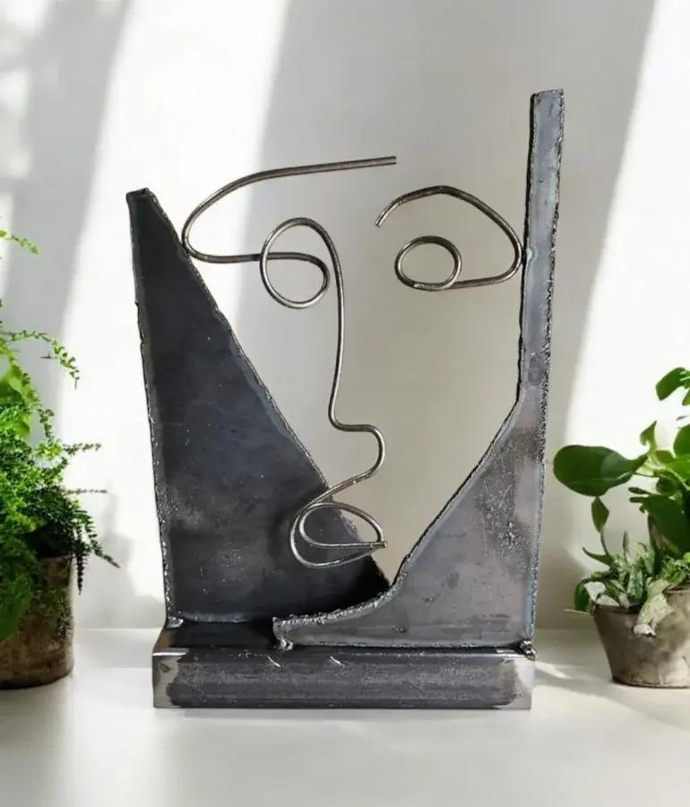 Abstract welded metal sculpture of a minimalist human face made from steel rods and plates, displayed on a white surface with green plants in the background.