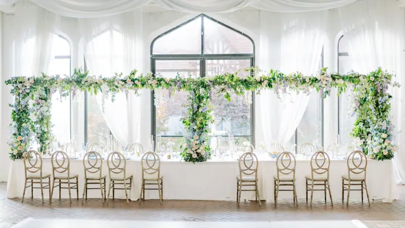 Indoor wedding canopy with greenery and flowers over a long head table and white draping.