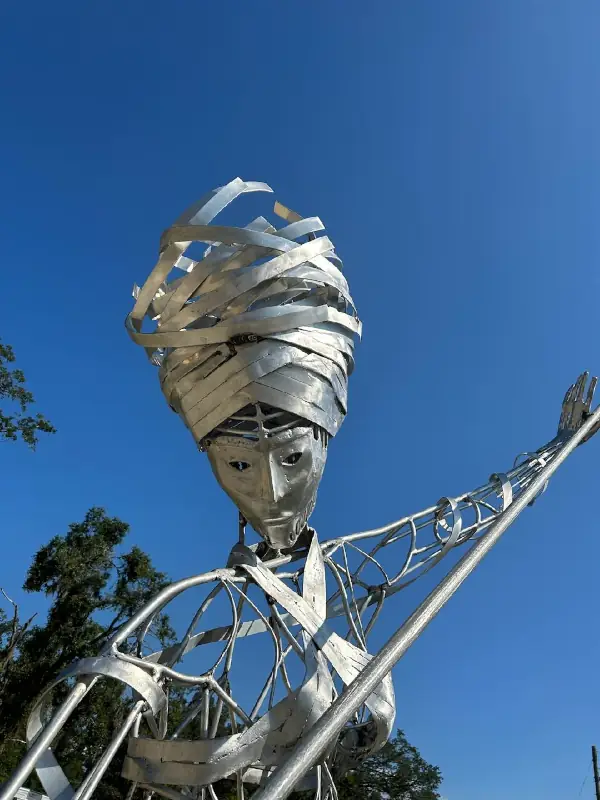 Large outdoor welded metal sculpture of a figure with a raised arm against a clear blue sky.